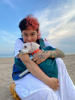 A photograph of a young person on a beach that kneels smiling in the sand while holding a small white dog with an orange collar. The person has gauged earrings, tattoos, orange hair,  and wears a a white skirt, a denim vest, and orange sandals.