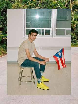 A photograph of a young person sitting on a stool and holding a Puerto Rican flag. They wear glasses, jeans, and yellow Nike, and sit on a small stool.