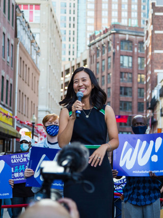 A photograph of Michelle Wu holding a microphone at a political rally in downtown Boston. She smiles as she stands in front of a crowd of supporters holding Michelle for Boston posters in a various languages.